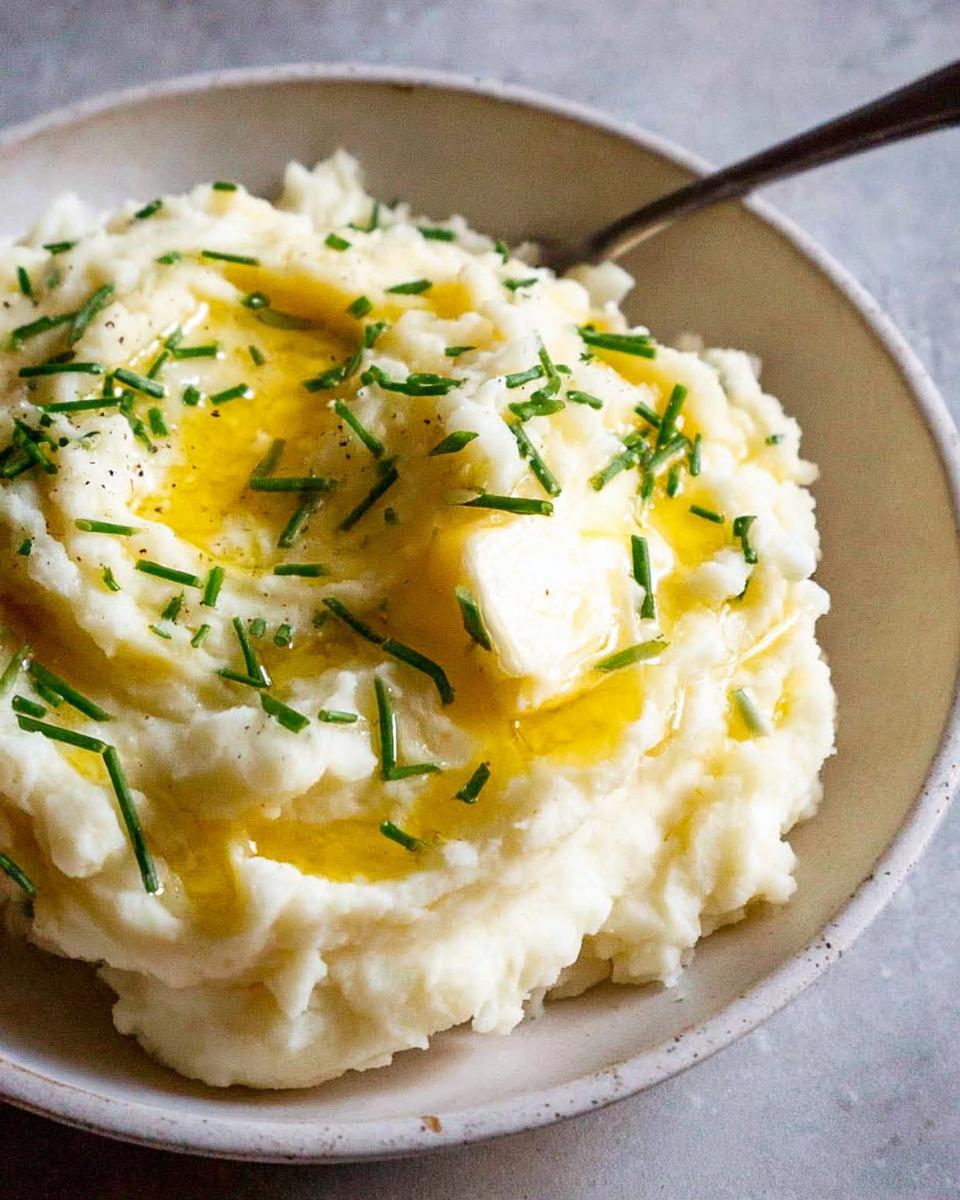 A close-up of creamy 12-Ingredient Mashed Potatoes topped with melted butter, chives, and black pepper.
