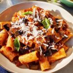 A close-up of rigatoni pasta with a rich tomato sauce, chunks of eggplant, and grated Parmesan cheese, garnished with fresh basil leaves.
