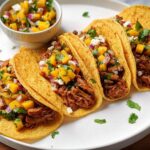 A plate of four corn tortilla tacos filled with shredded meat, mango salsa, crumbled cheese, and cilantro. Perfect for Taco Tuesday!