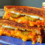 A close-up of a decadent 20-ingredient grilled cheese recipe, sliced in half to reveal melted cheese, pickles, and tomato.