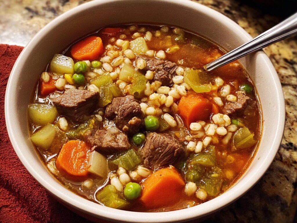 A close-up of a bowl of hearty beef barley soup, packed with tender beef chunks, carrots, peas, and barley.
