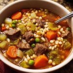 A close-up of a bowl of hearty beef barley soup, packed with tender beef chunks, carrots, peas, and barley.