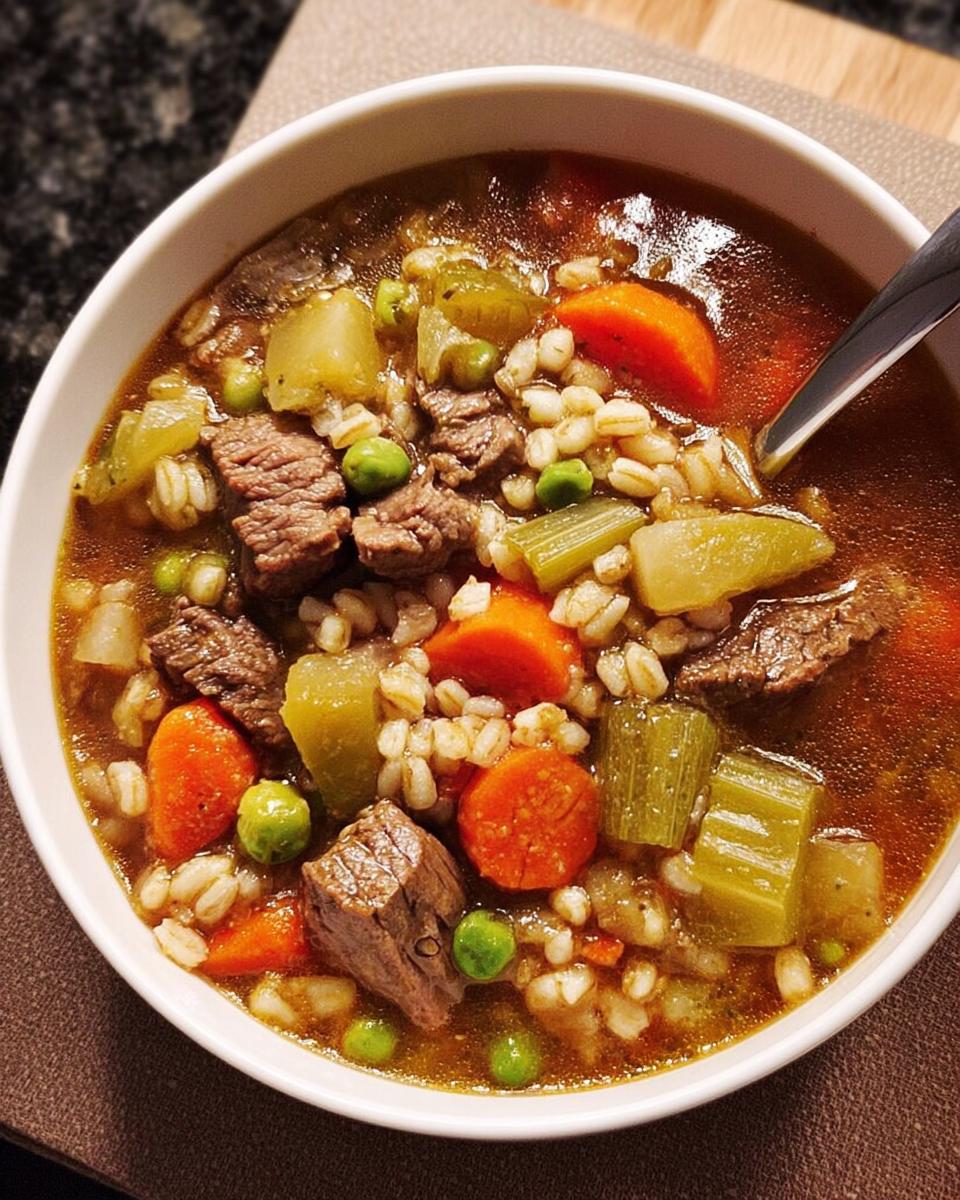 A close-up of a bowl of hearty beef and barley soup, featuring tender beef chunks, carrots, celery, peas, and barley.