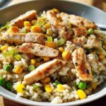 A close-up of a rice bowl filled with fluffy rice, grilled chicken pieces, corn, peas, and diced bell peppers.