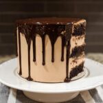 A tall, multi-layered chocolate cake with light brown frosting and rich chocolate ganache dripping down the sides.