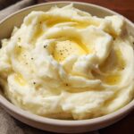 Close-up of fluffy mashed potatoes recipe topped with melted butter and cracked black pepper in a bowl.