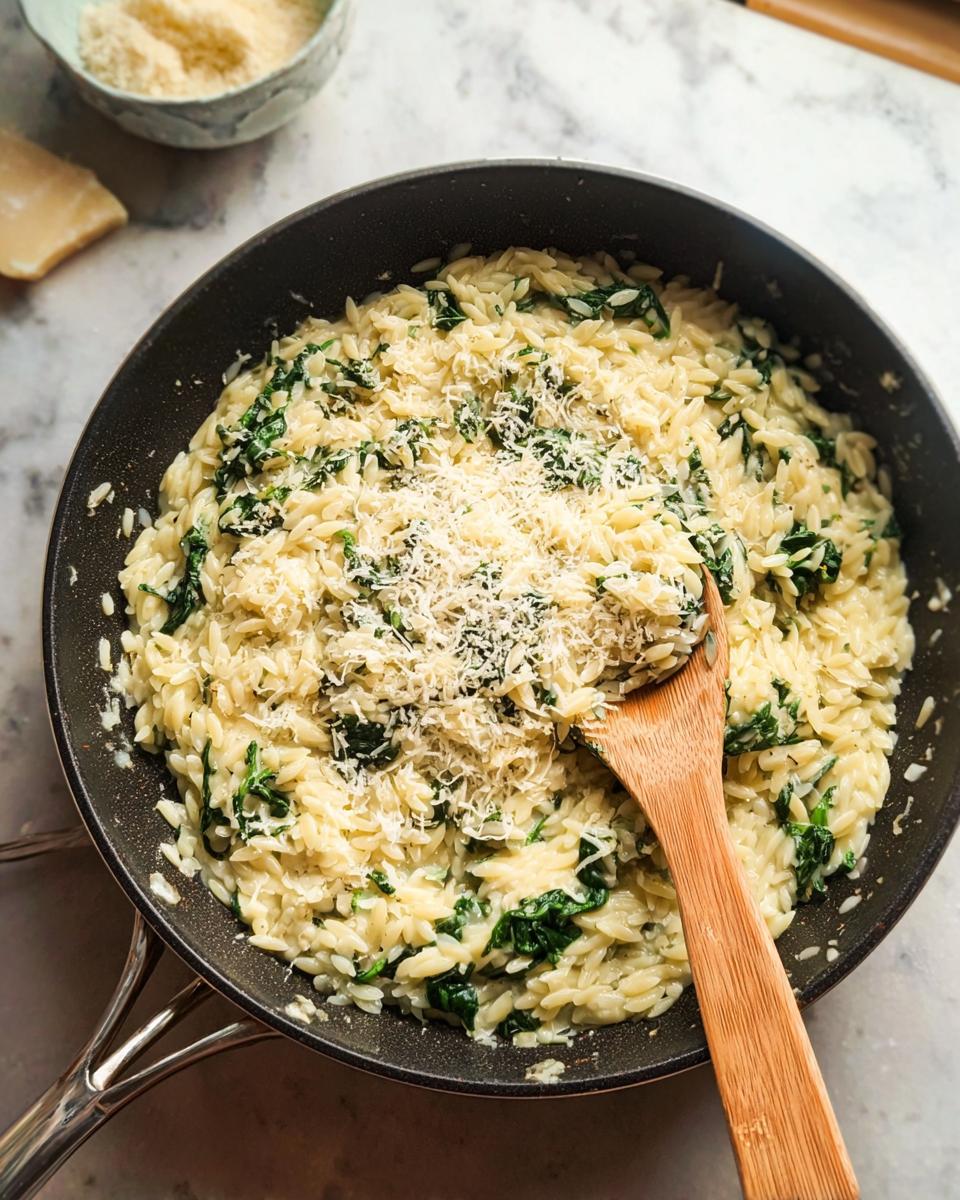 A close-up shot of creamy spinach orzo being stirred in a pan with a wooden spoon, topped with grated parmesan cheese.