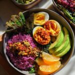 A vibrant bowl featuring purple rice, sliced hard-boiled eggs, avocado, orange slices, and crispy greens, part of an ultimate egg recipes guide.