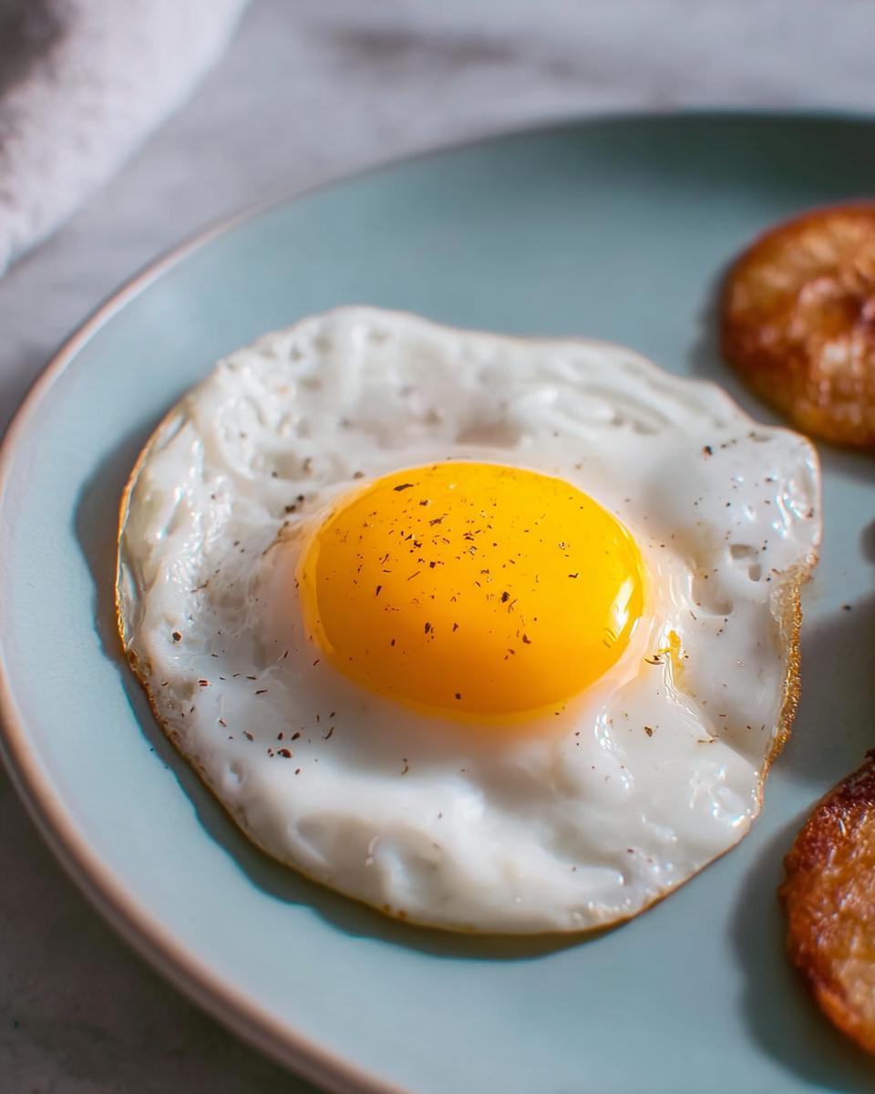 A perfectly fried egg with a bright yellow yolk, seasoned with pepper, on a blue plate, part of a delicious egg recipe.