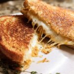 A perfectly grilled cheese sandwich, cut in half, with melted cheese oozing out. Features caramelized onions and herbs.