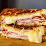 Close-up of a grilled ham and cheese sandwich, cut in half, with melted cheese oozing out.