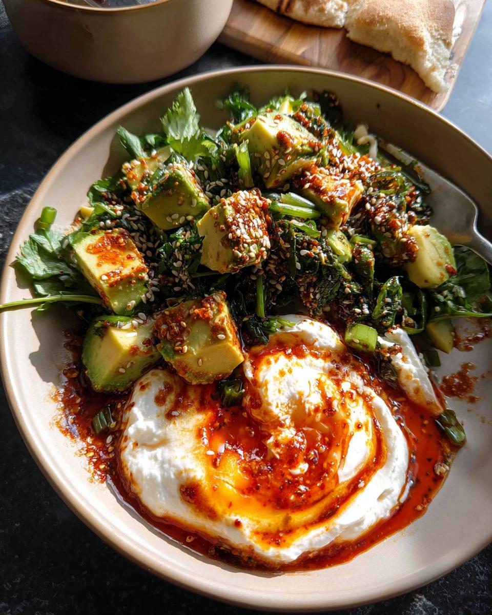 A healthy meal recipe featuring a bowl of fresh greens, diced avocado, and creamy yogurt with chili oil and sesame seeds.