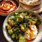 A healthy meal recipe featuring a bowl of diced avocado, herbs, and a poached egg with runny yolk, alongside a dip and bread.