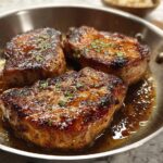 Three pan-seared pork chops in a skillet with a rich, glossy sauce and sprinkled herbs.