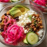 Close-up of a meal prep rice bowl featuring white rice, seasoned ground meat, black beans, crumbled feta, avocado slices, pickled red onions, and cucumber.