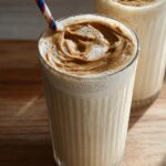 Close-up of a creamy peanut butter smoothie in a glass, topped with a swirl of peanut butter and a striped straw. Part of breakfast ideas.