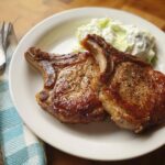 Two perfectly cooked pork chops served with a creamy side dish on a white plate.