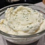 A close-up of a clear glass bowl filled with creamy, fluffy mashed potatoes, garnished with fresh parsley.