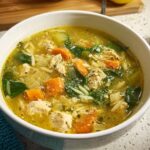 A steaming bowl of restaurant-style chicken noodle soup with carrots, celery, spinach, and orzo pasta.