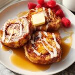 Close-up of restaurant-style cinnamon roll French toast drizzled with icing and syrup, served with raspberries.