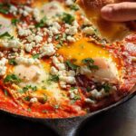 A hand dips bread into a skillet of restaurant-style shakshuka, a dish with eggs poached in tomato sauce and topped with feta cheese.