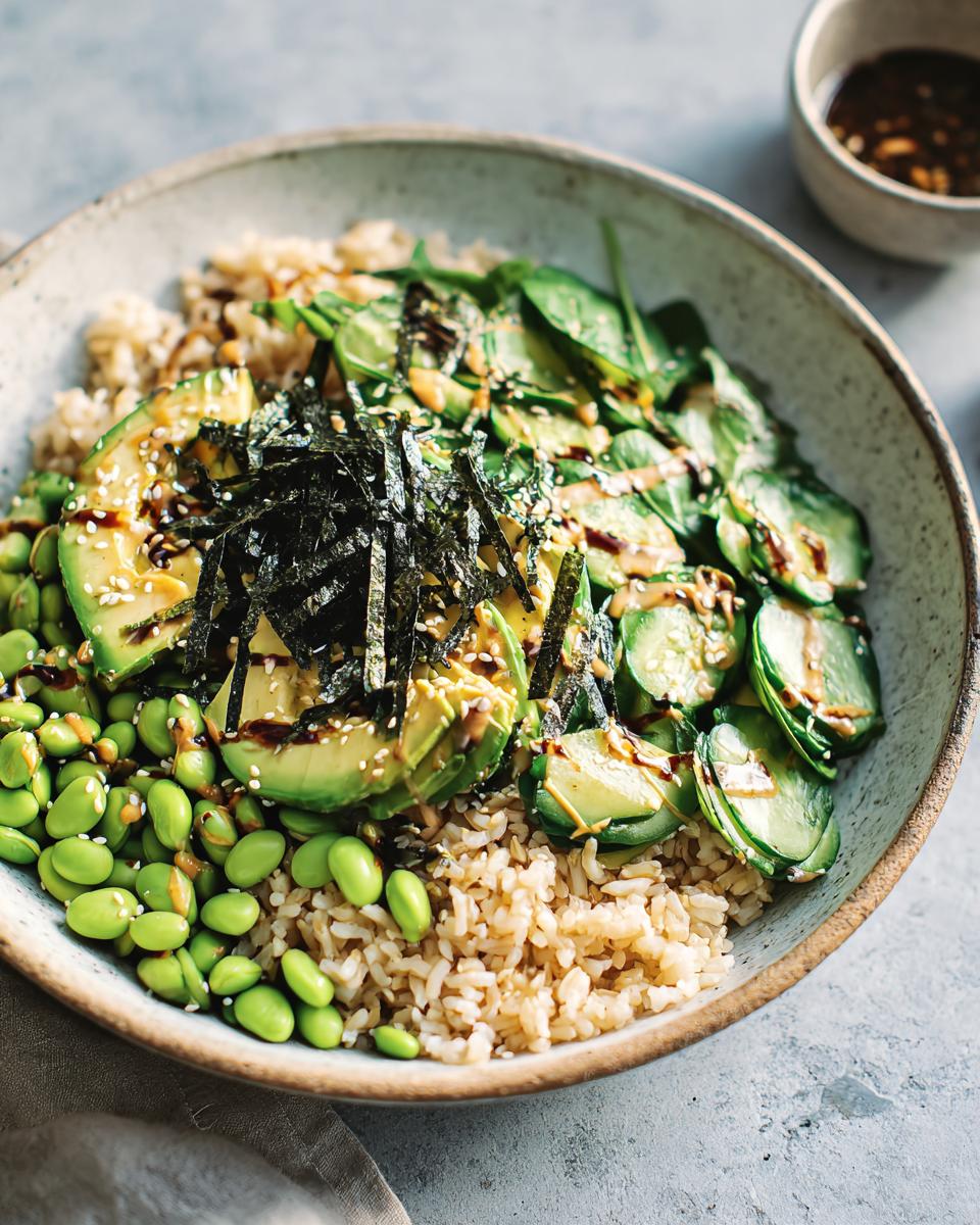 A vibrant and healthy rice bowl featuring brown rice, edamame, sliced avocado, cucumber, nori, and sesame seeds, drizzled with sauce.