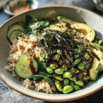 A vibrant and healthy rice bowl with brown rice, edamame, avocado, cucumber, spinach, and nori, drizzled with sauce.