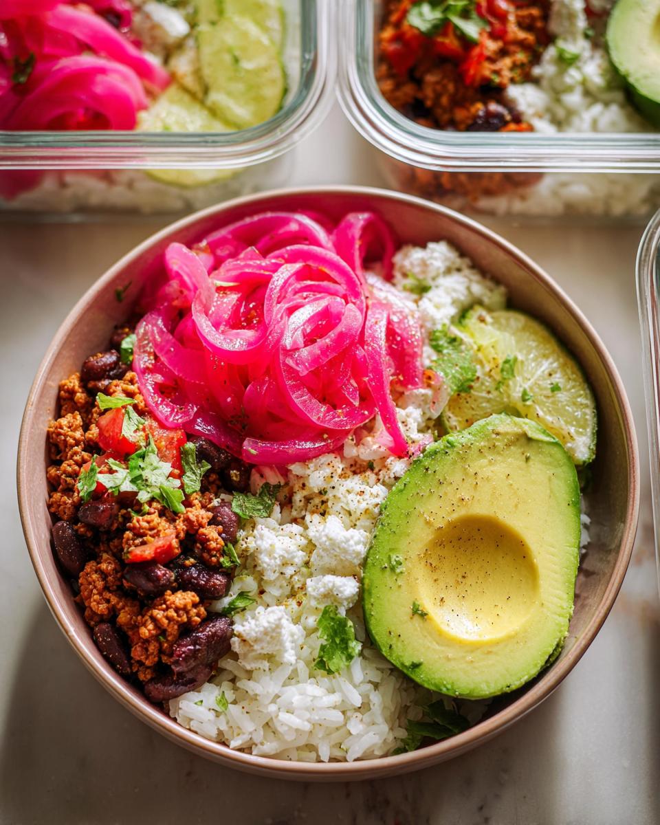 Close-up of a vibrant rice bowl with seasoned ground meat, black beans, white rice, crumbled cheese, pickled red onions, avocado, and lime.