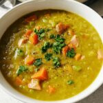 A close-up of a bowl of hearty split pea soup with chunks of ham, carrots, and celery, garnished with fresh parsley.
