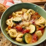 A bowl of tortellini vegetable soup with zucchini, tomatoes, and potatoes, topped with parmesan cheese.