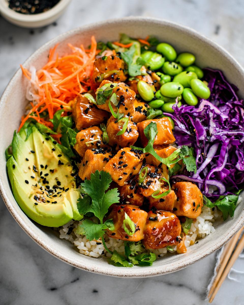 A vibrant bowl filled with rice, topped with glistening Bang Bang Salmon Bites, avocado, shredded carrots, edamame, and red cabbage.