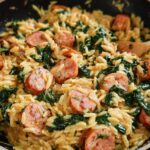 Close-up of Creamy Chicken Sausage Orzo with wilted spinach in a black skillet, garnished with a wooden spoon.
