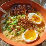 A close-up of a steaming bowl of Creamy Garlic Chicken Ramen, topped with sliced chicken, soft-boiled eggs, and green onions.