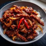 A close-up of a bowl of Crispy Chilli Beef Rice, featuring tender beef strips in a glossy sauce with onions and red chilies, served over white rice.
