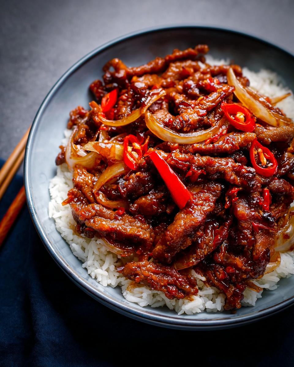 A close-up of a bowl of Crispy Chilli Beef Rice, featuring tender beef strips in a glossy sauce with onions and red chillies, served over white rice.