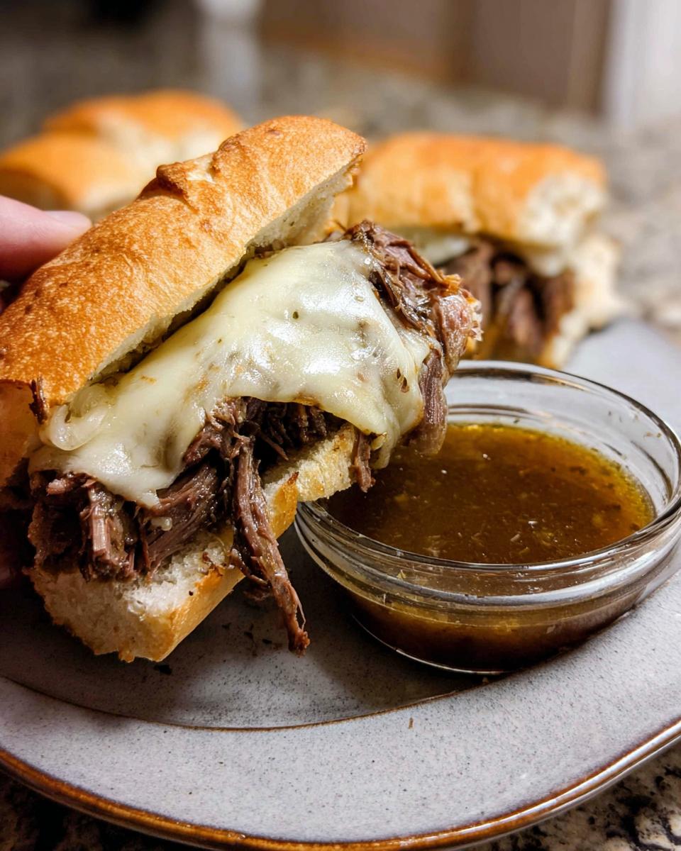 Close-up of a Crockpot French Dip Sandwich overflowing with shredded beef and melted cheese, next to a small bowl of au jus.