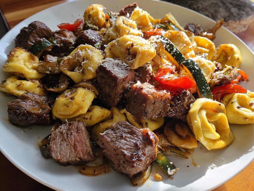 A close-up of a white plate filled with Garlic Steak Tortellini, featuring tender steak bites, pasta, zucchini, and tomatoes, topped with grated cheese.