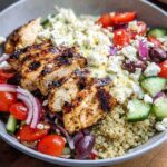 A close-up of a Greek Chicken Bowl with grilled chicken, quinoa, tomatoes, cucumbers, olives, red onion, and feta cheese.