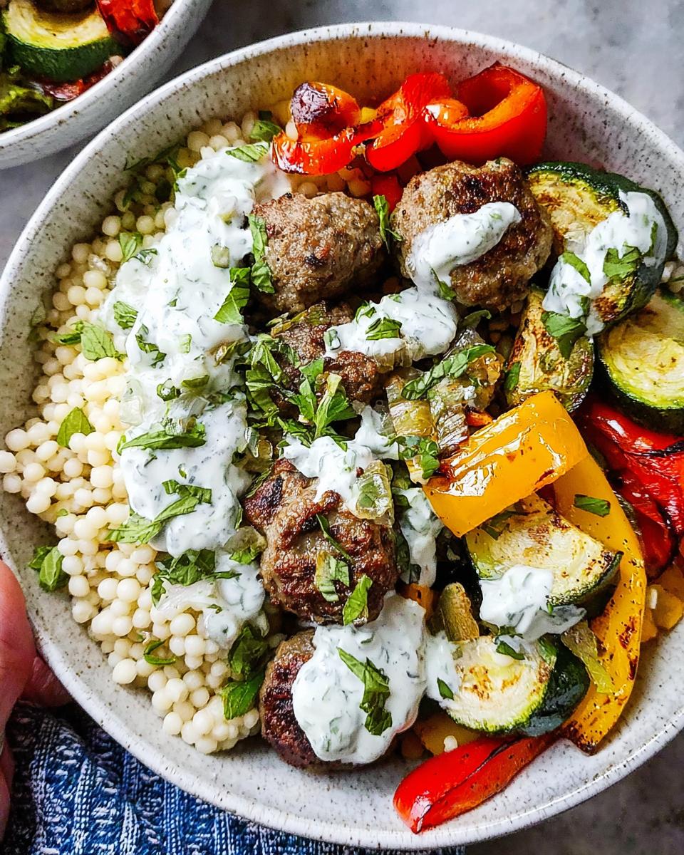 A delicious Greek meatball bowl filled with couscous, meatballs, roasted bell peppers, zucchini, and drizzled with a creamy sauce.