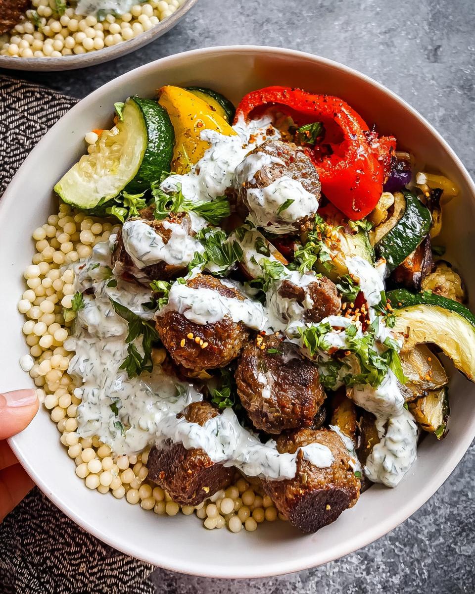 A close-up of a Greek Meatball Bowl Recipe featuring juicy meatballs, roasted vegetables, and couscous, drizzled with creamy sauce.