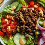 A close-up of a High-Protein Cheeseburger Bowl with seasoned ground beef, lettuce, tomatoes, cucumbers, and red onion, drizzled with sauce.