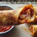 A close-up of a HIGH PROTEIN Pepperoni Pizza Roll, showing melted cheese, pepperoni, and marinara sauce, with more rolls in the background.