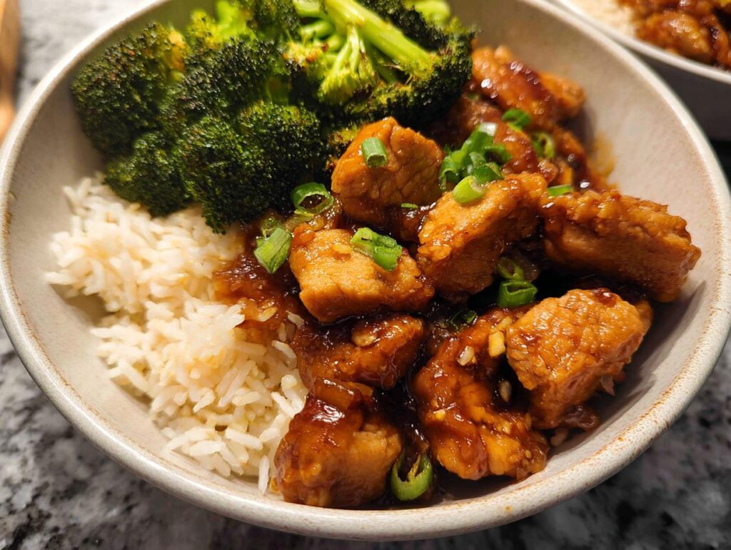 A delicious Honey Garlic Pork Rice Bowl featuring tender pork pieces in a glossy sauce, fluffy white rice, and steamed broccoli florets, garnished with green onions.