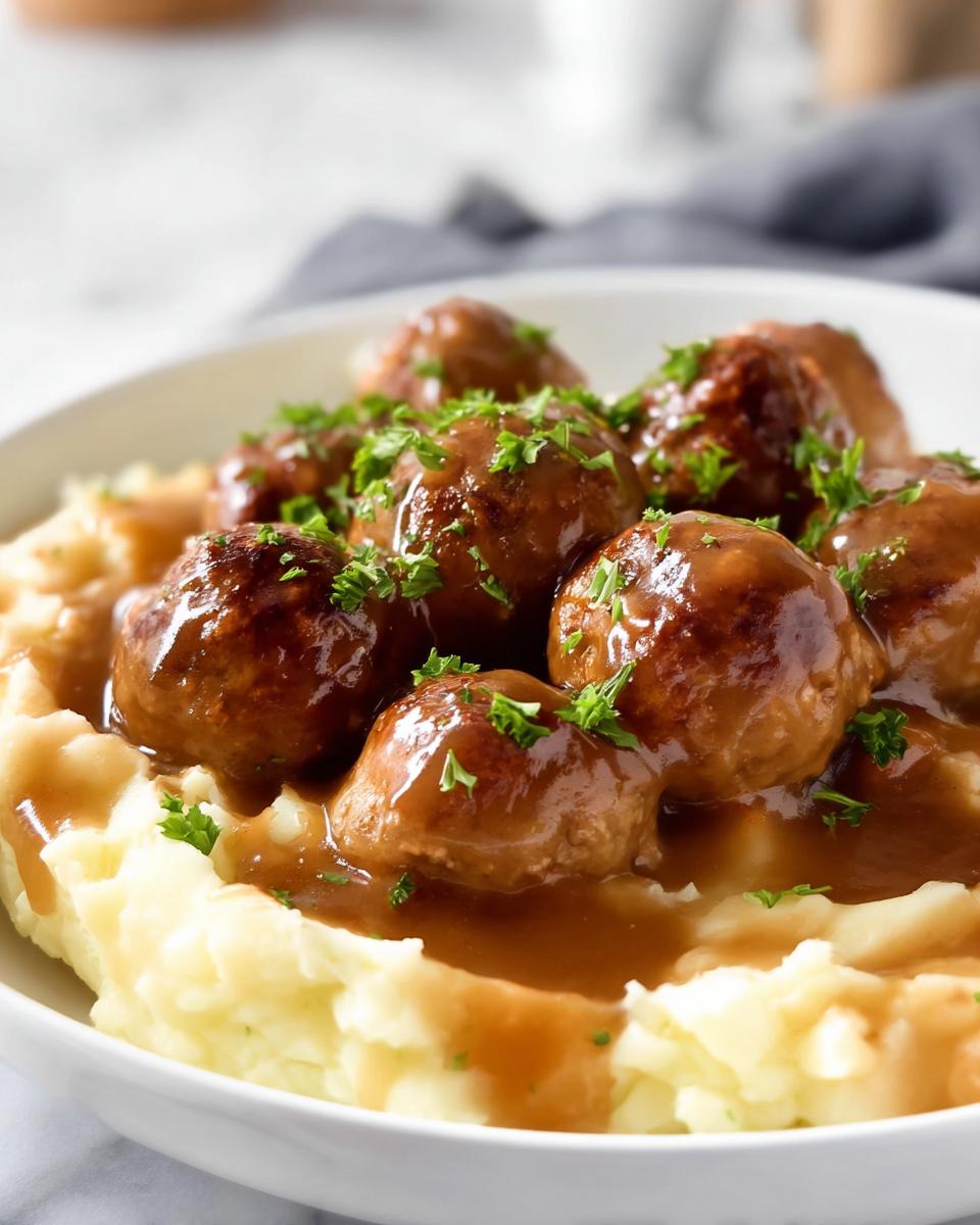A close-up of Salisbury Steak Meatballs smothered in gravy, served over creamy Garlic Herb Mashed Potatoes and garnished with parsley.