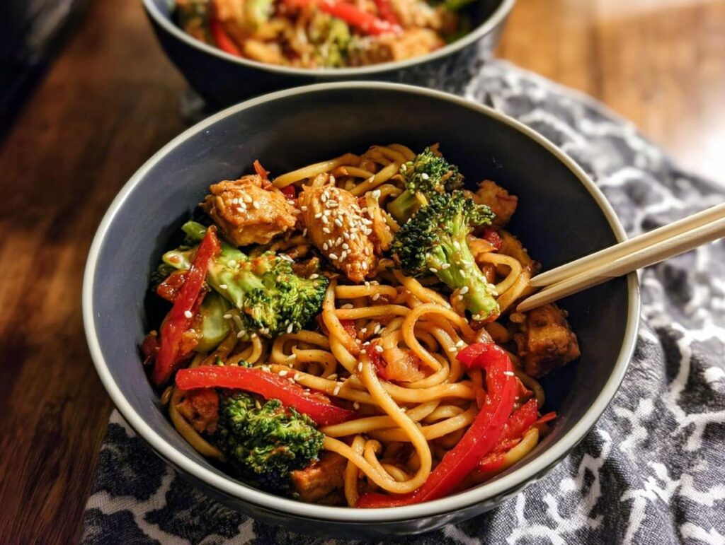 A close-up of a bowl of Spicy Garlic Chicken and Broccoli Noodle Bowls, featuring noodles, chicken, broccoli, and red peppers, topped with sesame seeds.