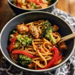 A close-up of a bowl of Spicy Garlic Chicken and Broccoli Noodle Bowls, featuring noodles, chicken, broccoli, and red peppers, topped with sesame seeds.