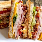 Close-up of a delicious Breakfast Club Sandwich with layers of turkey, ham, bacon, cheese, lettuce, and tomato on toasted bread.