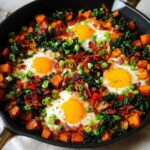 A cast iron skillet filled with BREAKFAST SWEET POTATO hash, topped with fried eggs, bacon, and green onions.