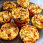 A plate of golden-brown Cheesy Muffin Tin Cottage Cheese Egg Bites, some with visible bits of red pepper and green herbs.