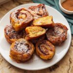 A plate filled with golden brown Cinnamon Roll French Toast Bites, dusted with powdered sugar.